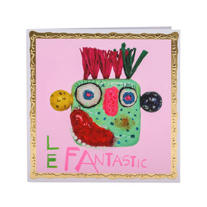 A brightly colored greetings card featuring a mask made by Arthouse Unlimited artists, of a playful, cartoon-like face with exaggerated features. The background is a light pink, and the card is outlined with a gold border. The text "LE FANTASTIC" is written below the face in a playful font.