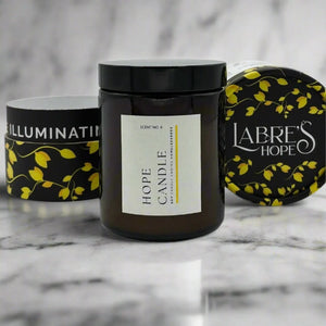 Three candles with black glass jars and white labels with yellow vines sit on a marble surface.