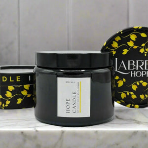 Three candles with black glass jars and white labels with yellow vines sit on a marble surface.