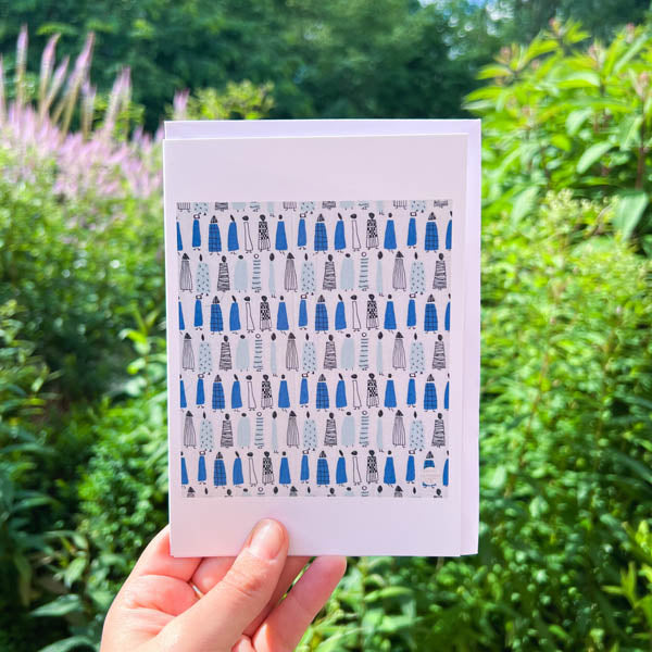 A person holding a greetings card in front of a garden backdrop. The print is a reproduction of Lucienne Day's Sequence design. Repeat pattern of women in blue coats