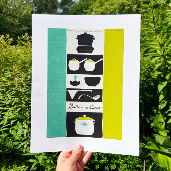 A person holding up a print in front of a garden backdrop, the print has an image of Batterie de Cuisine by Lucienne Day. A green, black and white print with Kitchenware at the centre.
