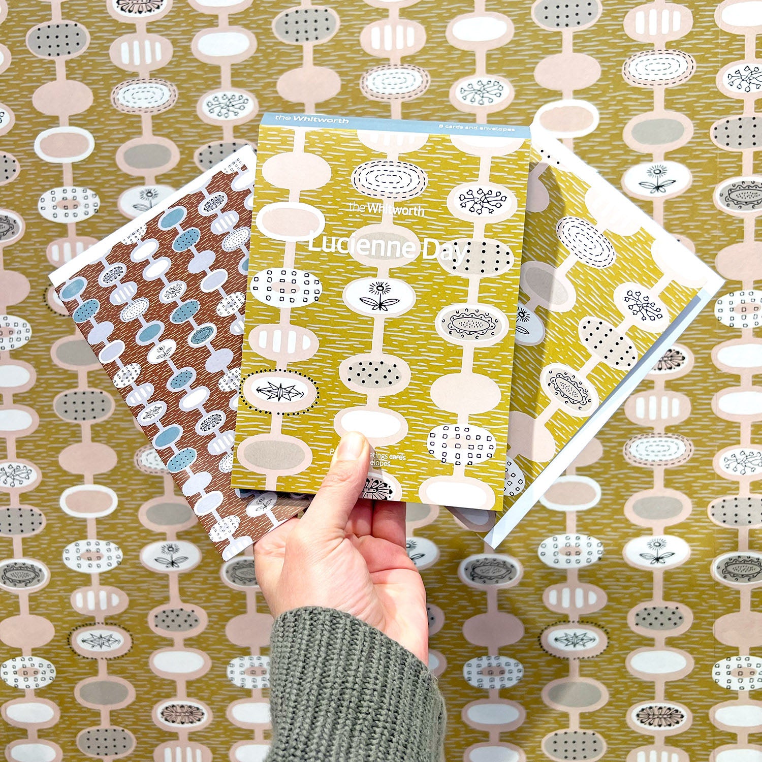 A person holding up a set of greetings cards featuring a yellow, pink and white repeat pattern design by Lucienne Day. They are holding it up in front of a wallpaper of the same design in the Whitworth Shop.