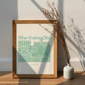 Framed print of a green line illustration of the Manchester skyline.  The print is displayed on a wooden floor with a vase of dried flowers beside it and sunlight streaming in from a window.