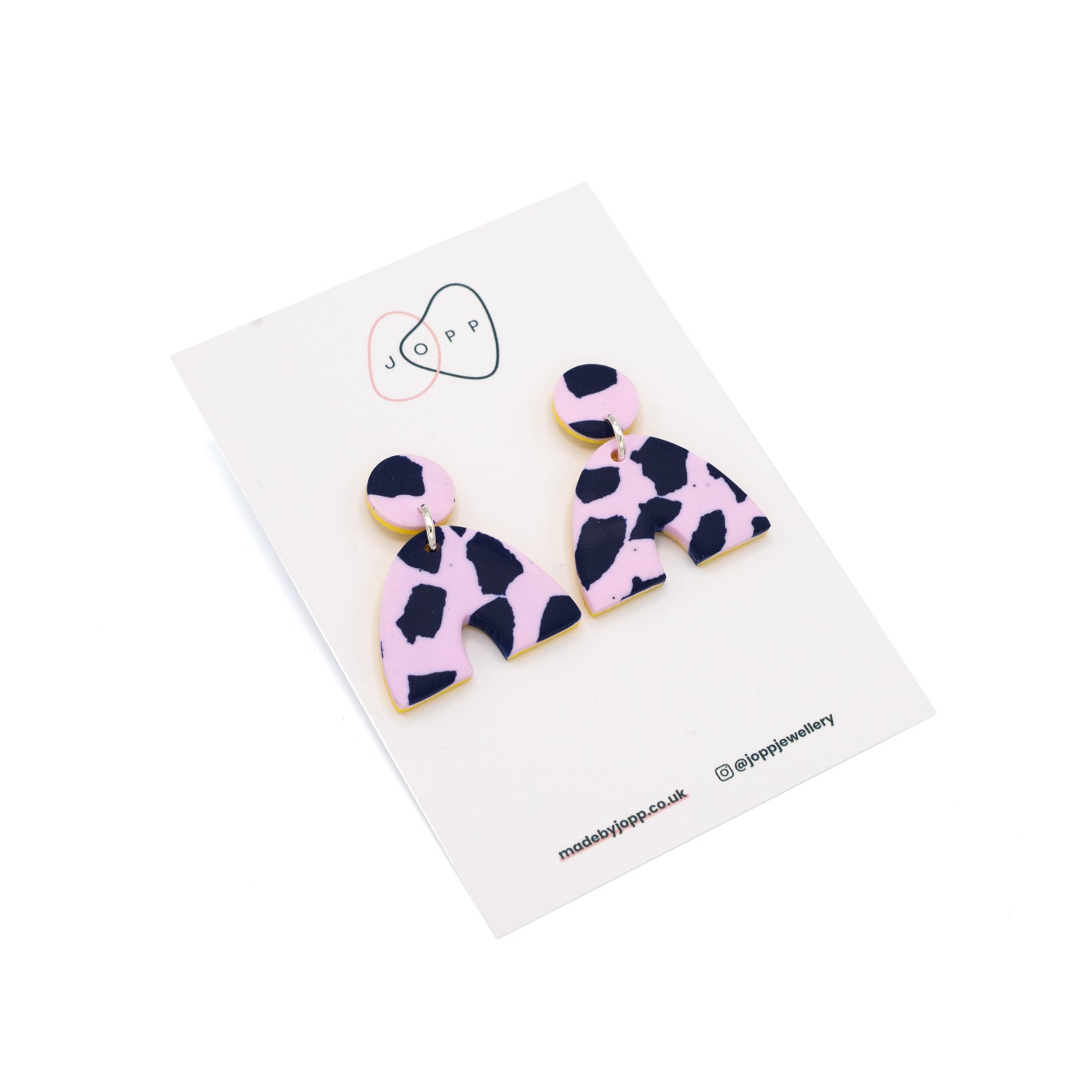 Earrings in a horseshoe shape photographed on top of backing card with Jopp branding. The earrings have a pink and navy cow print pattern.