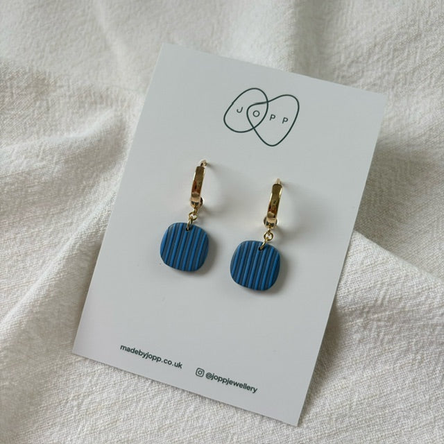 A pair of blue striped polymer clay earrings by JOPP Jewellery. The square earrings hang from gold-toned hoops and are displayed on a white card with the JOPP logo and website information.