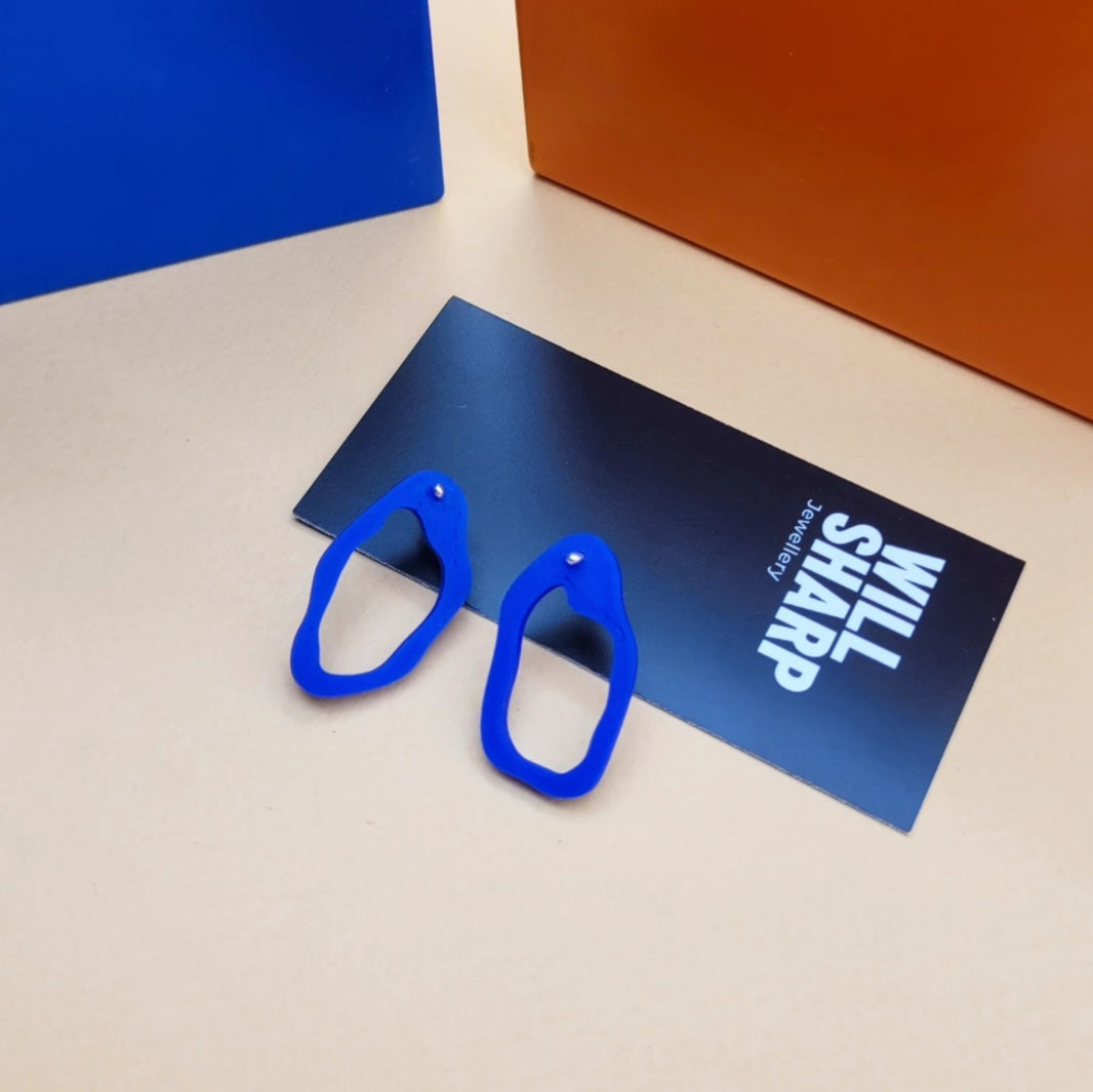Irregular shaped blue 3D printed earrings on branded backing card on a beige surface.