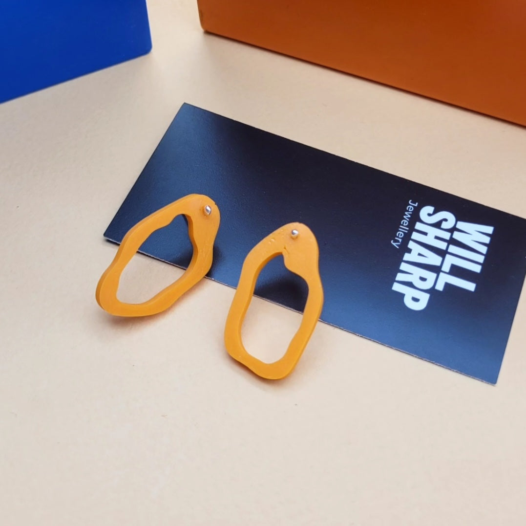 Irregular shaped orange 3D printed earrings on branded backing card on a beige surface.