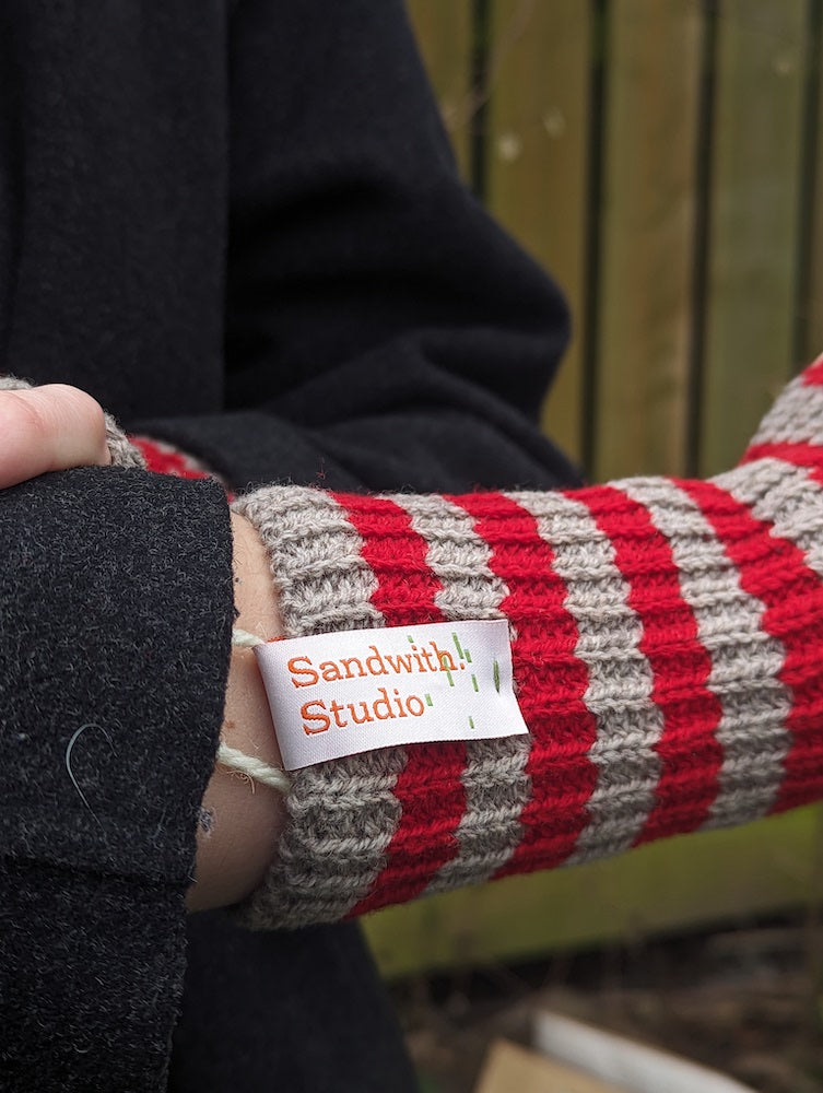 A close up of someone wearing red and grey striped fingerless gloves. Sandwith Studio label on gloves.