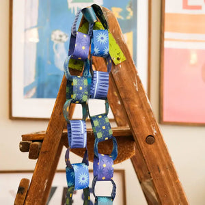 A photo of a colourful paper chain hanging from a wooden stepladder. The chain is made of blue, yellow, and green paper strips with patterns of stars and squares.