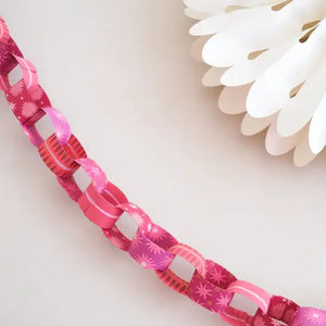 A close-up photo of a pink paper chain on a white background. The chain has a gradient of pink shades and features a pattern of stars. A white paper fan is partially visible in the background.