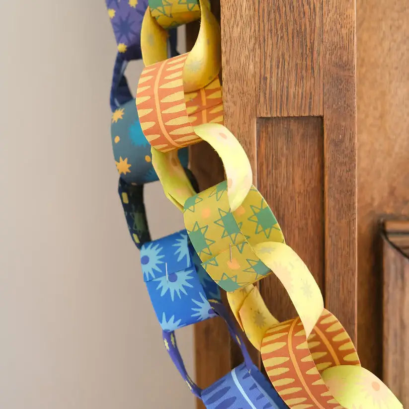 A close-up photo of a paper chain hanging from a wooden door frame. The chain is made of colourful paper strips with patterns, including stars, stripes, and dots.