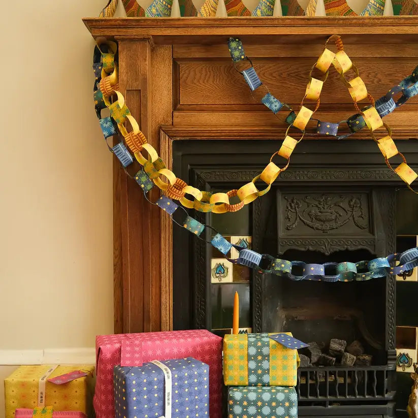 A photo of a decorated fireplace mantel. A long paper chain with alternating yellow and blue links hangs above the fireplace. There are also wrapped gifts and candles on the floor below.