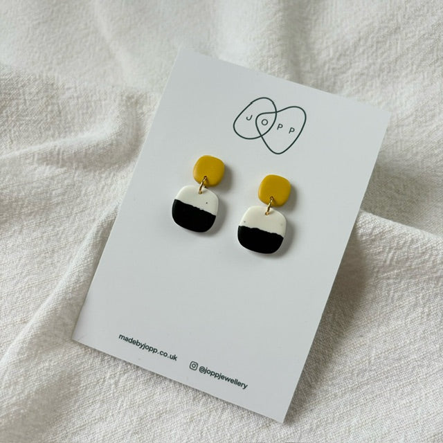 A pair of handmade polymer clay earrings by JOPP Jewellery. The earrings feature a square shape with a black and white gradient design and a small gold-colored top section. They are displayed on a white card with the JOPP logo and website information.