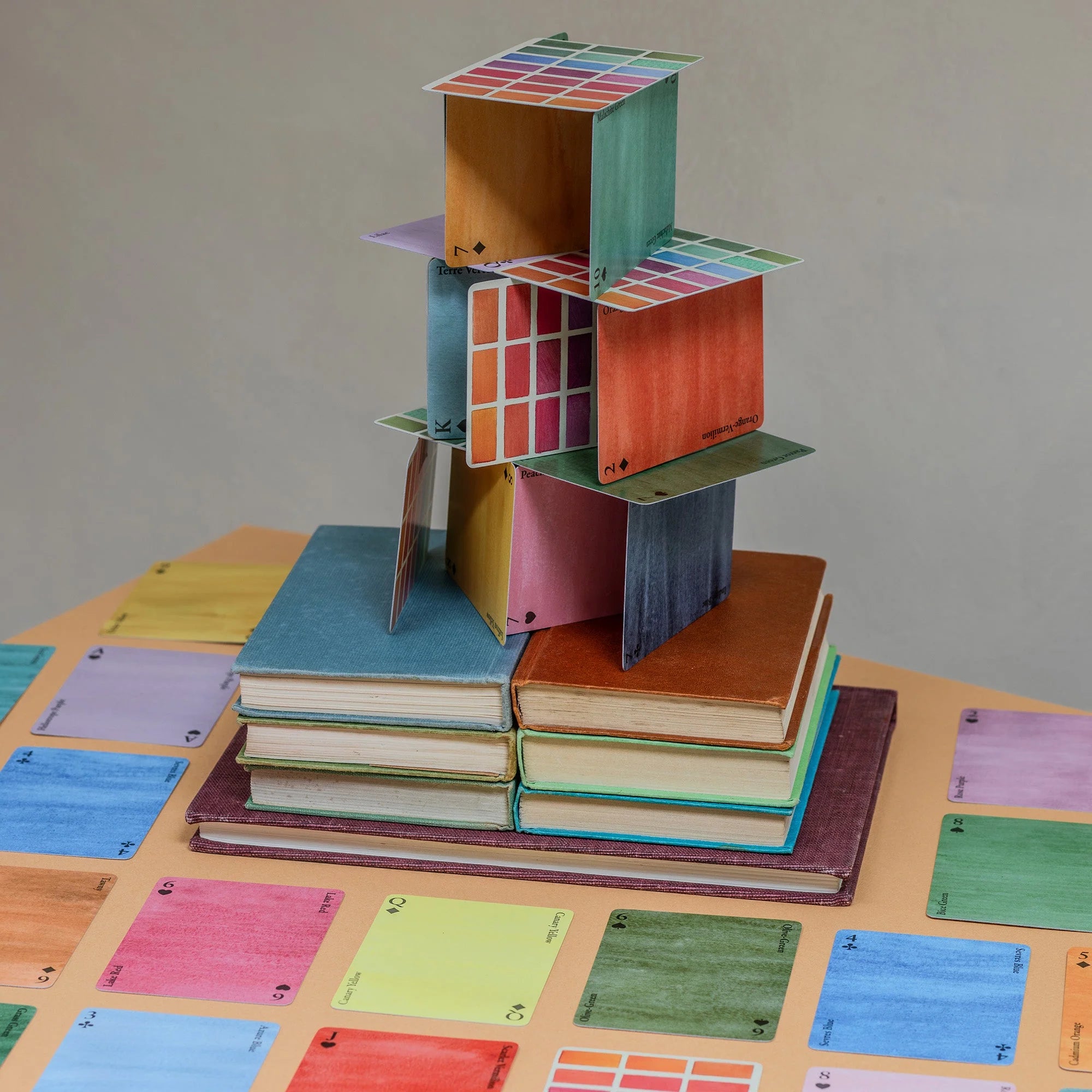 A stack of books with a tower of playing cards built on top. The playing cards feature different colours and names from the "Nomenclature of Colours" series, while the books are in various colours and sizes. The background shows additional playing cards and books scattered across the table.