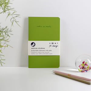 Green journal leant against a white wall with pencils and greenery coming in from outside the frame.
