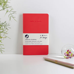 Red journal leant against a white wall with pencils and greenery coming in from outside the frame.