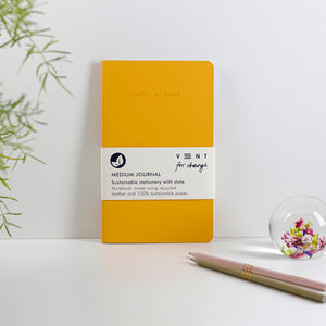 Yellow journal leant against a white wall with pencils and greenery coming in from outside the frame.