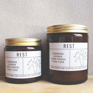 Both sizes of the rest candle standing next to each other with the smaller to the left. The candles are of brown glass with gold metal lids. Simple paper branding stickers on the front in white and black text.