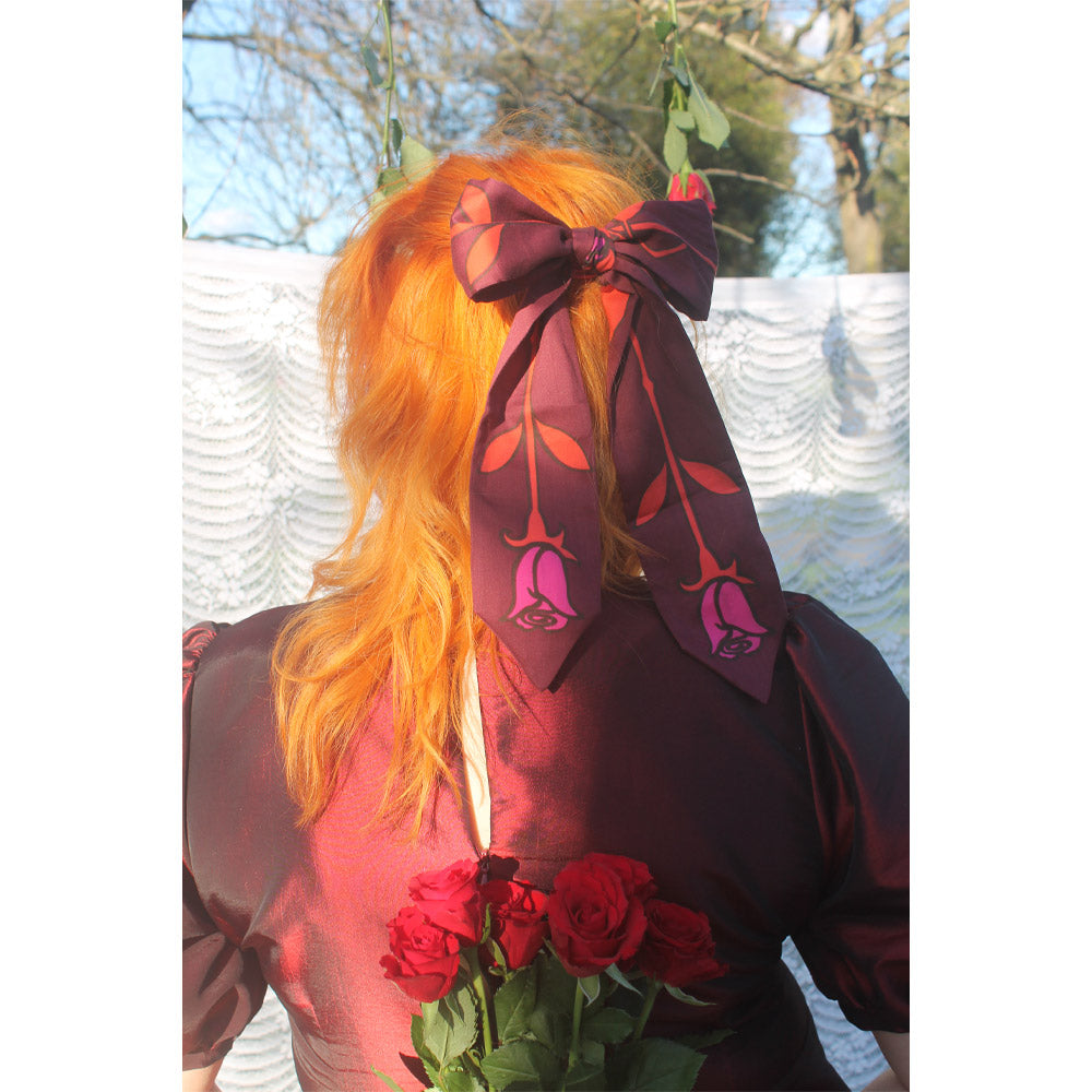 Person with bright orange hair wearing a large purple and red scarf tied in a bow around their hair, standing outdoors with a blurred background.