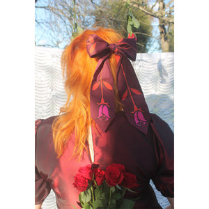 Person with bright orange hair wearing a large purple and red scarf tied in a bow around their hair, standing outdoors with a blurred background.