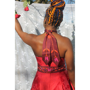 Person in a red dress with floral patterns standing against a white textured wall, wearing a red scarf with rose pattern