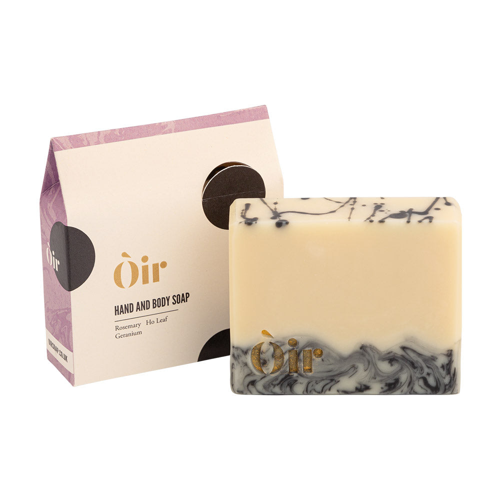 A bar of Oir Hand and Body Soap in a Rosemary, Ho Leaf, and Geranium scent sits on a white background next to its packaging.