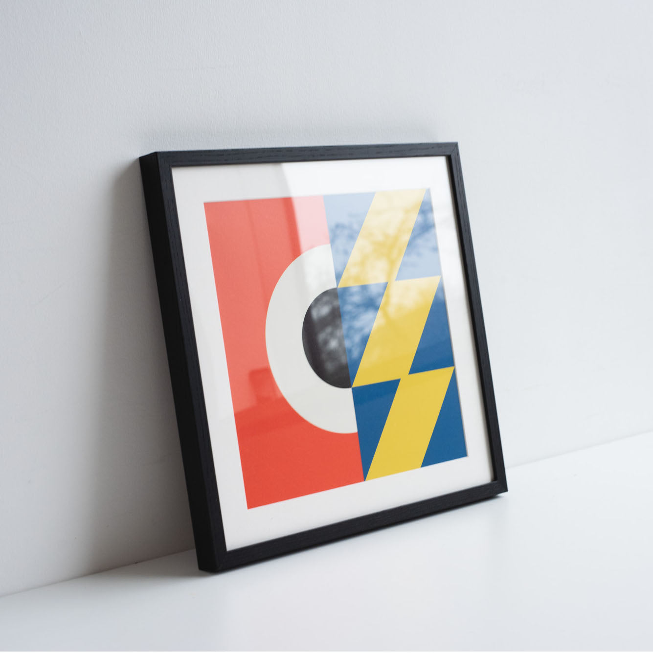 Square geometric digital illustration by Ruth Tuck in a black frame rested against a white wall, in red, white, black, blue and yellow.