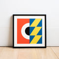 Square geometric digital illustration by Ruth Tuck in a black frame rested against a white wall, in red, white, black, blue and yellow.