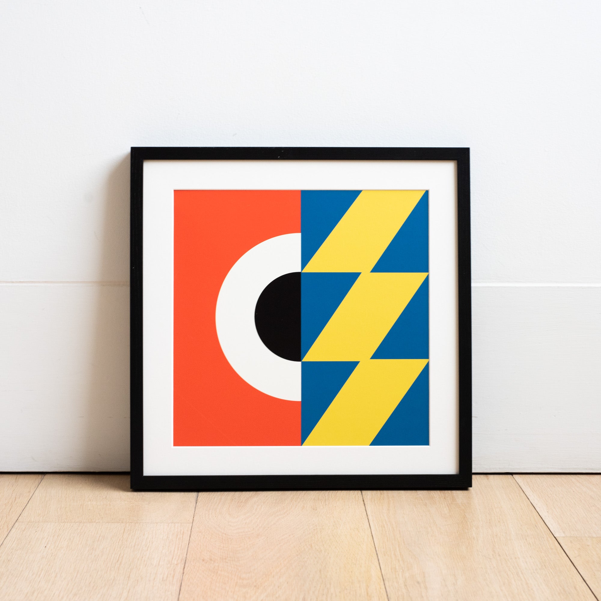 Square geometric digital illustration by Ruth Tuck in a black frame rested against a white wall, in red, white, black, blue and yellow.