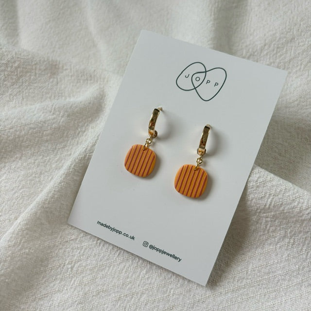 A pair of orange and red striped polymer clay earrings by JOPP Jewellery. The square earrings hang from gold-toned hoops and are displayed on a white card with the JOPP logo and website information.