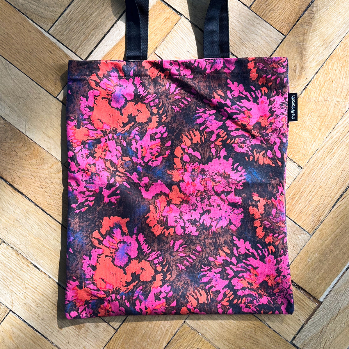 The image shows a tote bag with a vibrant floral pattern in shades of pink, orange, and black. The tote bag has black handles.