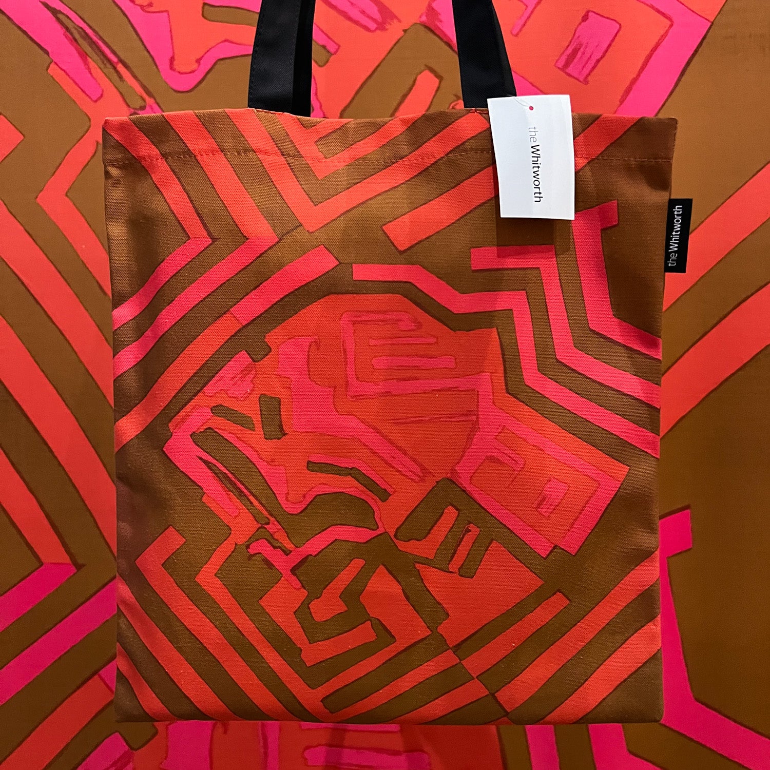  A person holding a tote bag with a black handle featuring Shirley Craven's sixty three design, in front of the textile design in the Whitworth's exhibition space. The tea towel has a white label attached with Whitworth's logo.