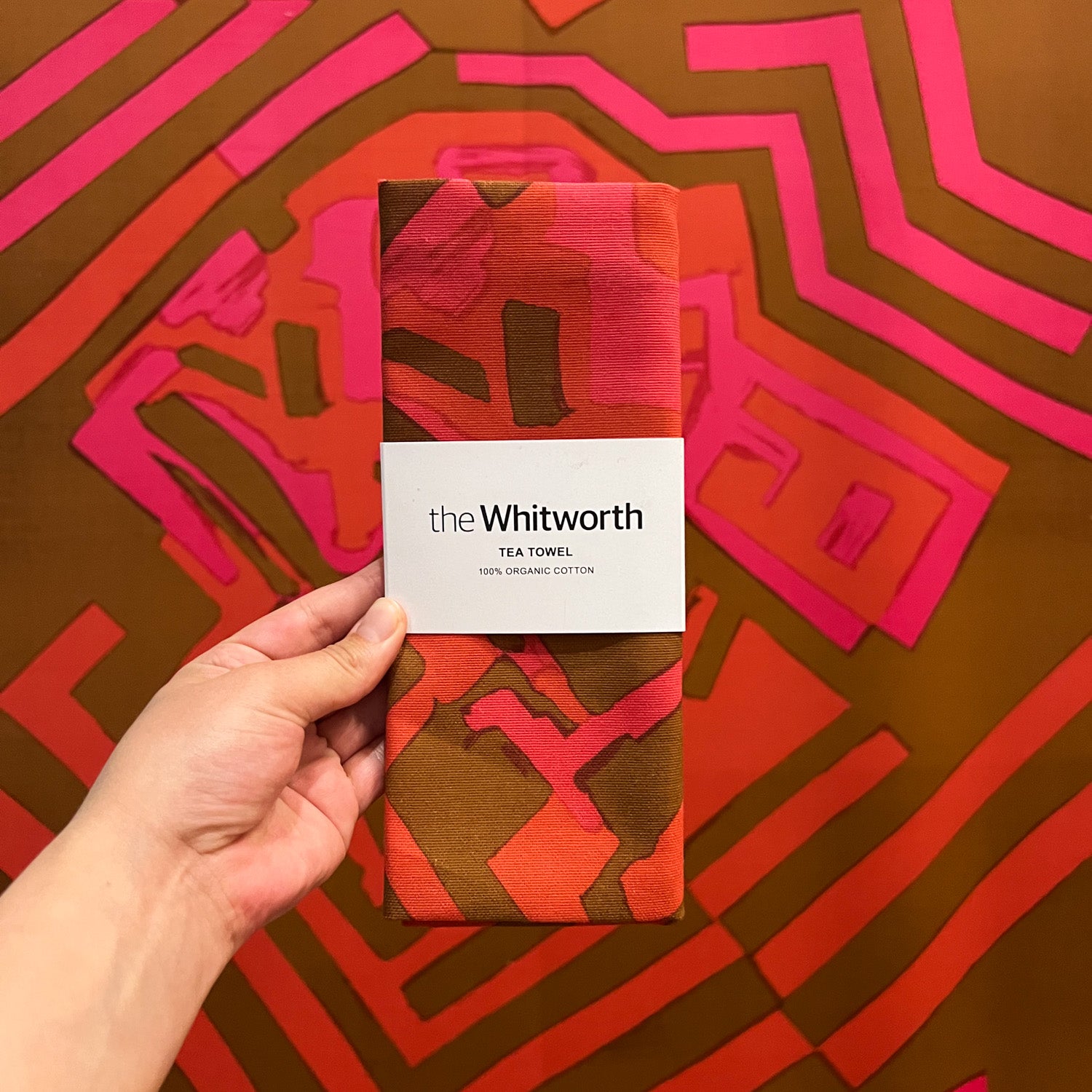 A person holding a tea towel featuring Shirley Craven's sixty three design, in front of the textile design in the Whitworth's exhibition space. The tea towel has a white belly band wrapped around it with the Whitworth's logo.