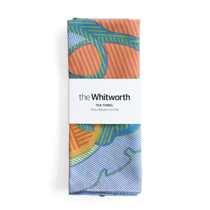 Tea towel with a vibrant pattern featuring blue, orange, and pink shapes. This pattern is a detail of a design by Shirley Craven. Belly band around the tea towel with the Whitworth logo.