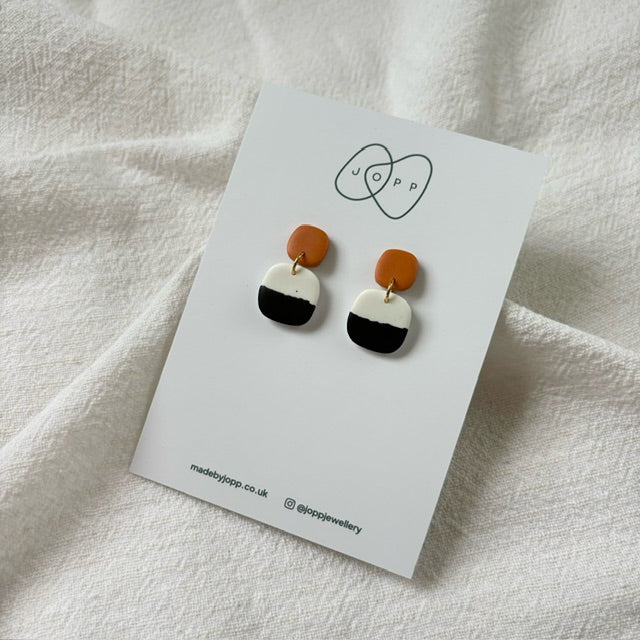 A pair of handmade polymer clay earrings by JOPP Jewellery. The earrings feature a square shape with a black and white gradient design and a small orange top section. They are displayed on a white card with the JOPP logo and website information.