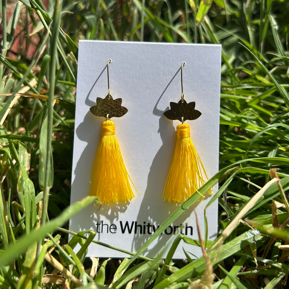 Yellow tassel earrings on a card with 'the Whitney' branding, placed on grass.