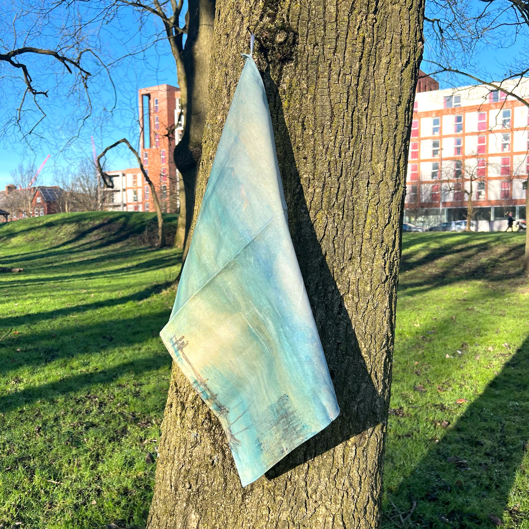 Tea Towel featuring JMW Turner watercolour titled Moonlight on Lake Lucerne with the Rigi in the Distance. The tea towel is hanging on a tree in The Whitworth Park.