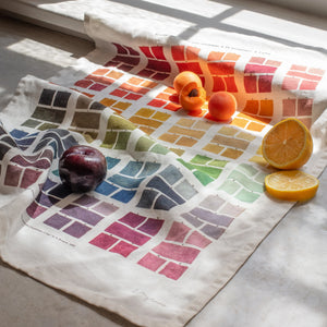 Tea towel featuring a grid of watercolour swatches in various colours, displayed on a windowsill with a plum and sliced oranges placed on top. 