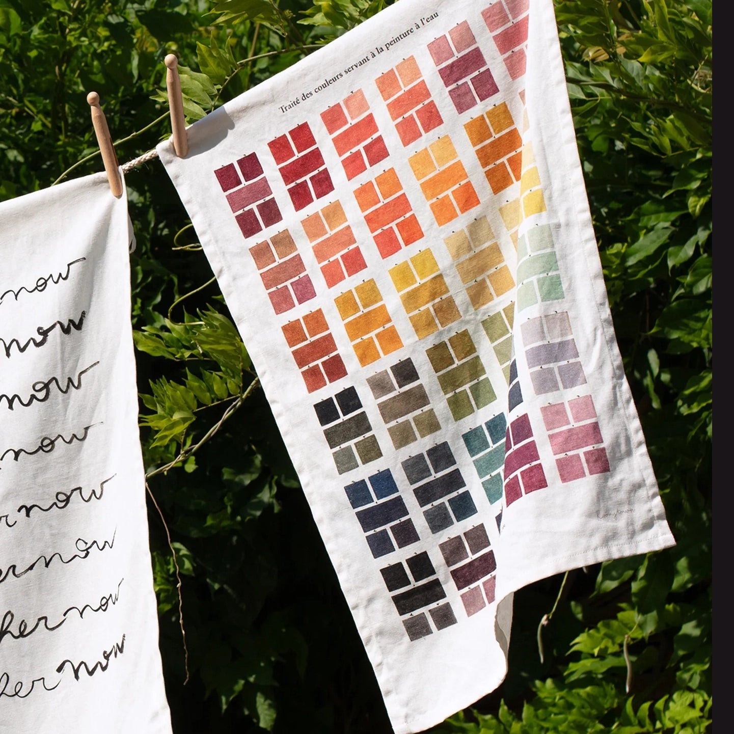 Tea towel featuring a grid of watercolour swatches in various colours, hung on a washing line in front of some greenery. 