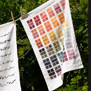 Tea towel featuring a grid of watercolour swatches in various colours, hung on a washing line in front of some greenery. 