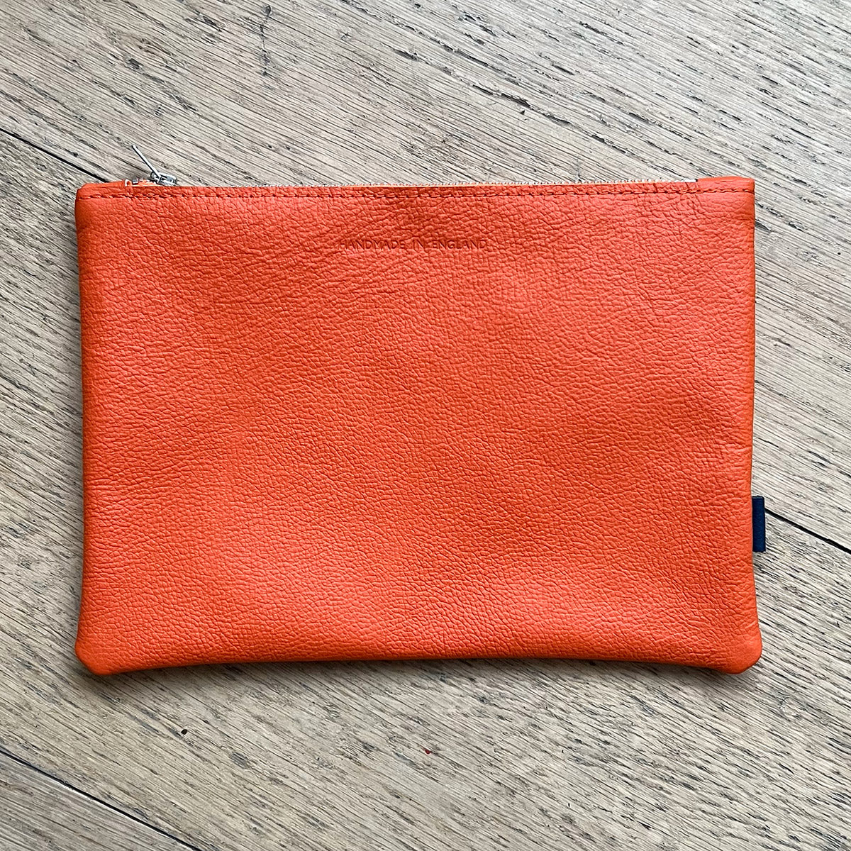 Rectangular orange leather pouch on a wooden surface. "Handmade in England" is stamped at the top centre.