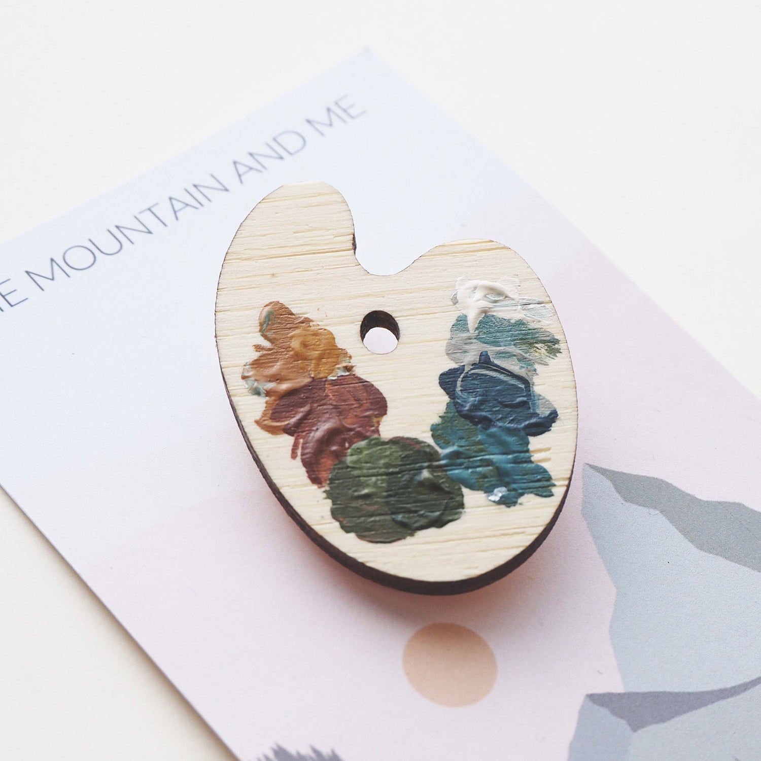 Brooch in the shape of a paint palette placed on a Mountain and Me Branded backing card