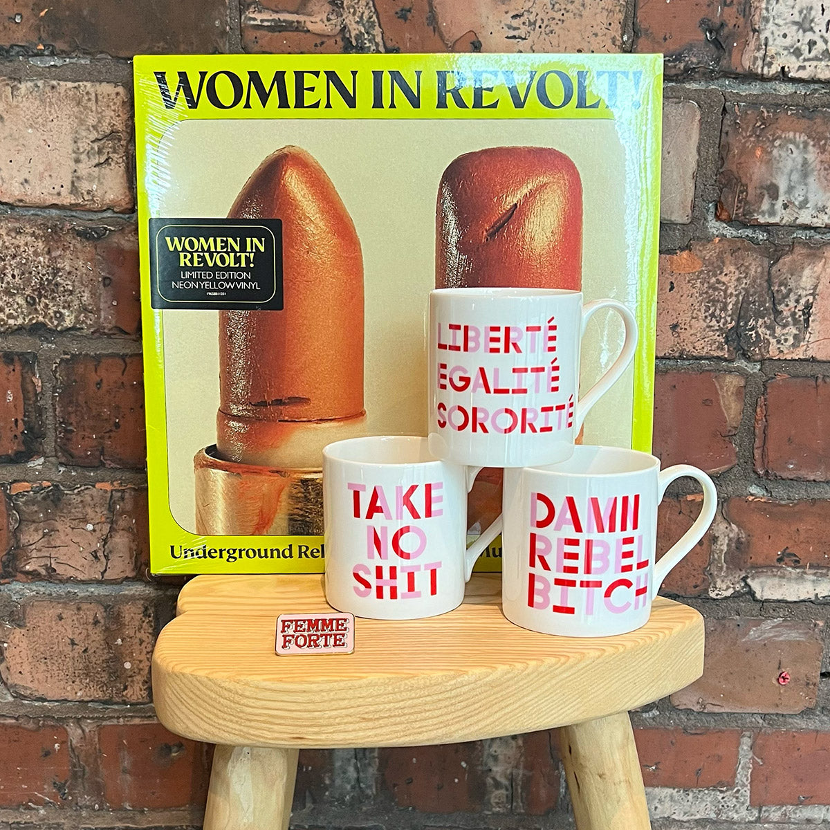 A yellow vinyl sleeve for the Women in Revolt record printed in front of a brick wall on a stool. There are three mugs stacked in front of it.