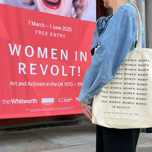 A person wearing a Women in Revolt tote bag, stood in front of the exhibition banner at The Whitworth