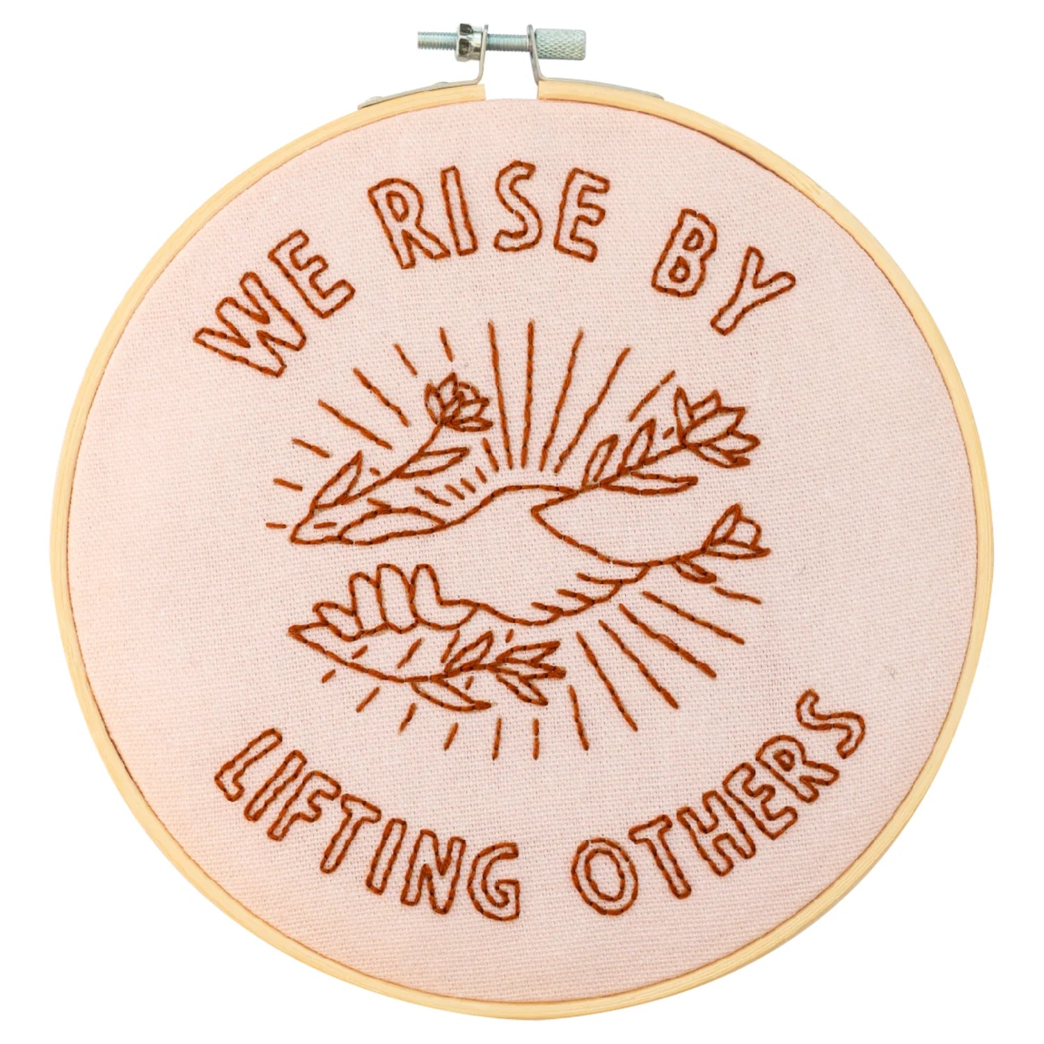 A round embroidery hoop with a pink fabric background and a brown thread design. The design shows two hands reaching upwards, with a sun-like symbol and leaves in the background. The words "WE RISE BY LIFTING OTHERS" are embroidered around the design.