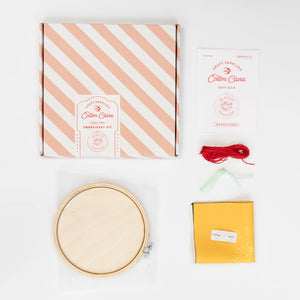 A cross-stitch kit laid out on a white background. It includes a wooden hoop, pink fabric, colourful thread, instructions, and a cardboard box with the brand name.