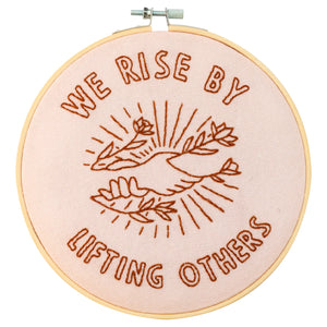 A round embroidery hoop with a pink fabric background and a brown thread design. The design shows two hands reaching upwards, with a sun-like symbol and leaves in the background. The words "WE RISE BY LIFTING OTHERS" are embroidered around the design.