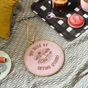 A round embroidery hoop with a pink fabric background and a brown thread design, hanging from a yellow ribbon. The design shows two hands reaching upwards, with a sun-like symbol and leaves in the background. The words "WE RISE BY LIFTING OTHERS" are embroidered around the design. The hoop is resting on a blanket with other craft supplies visible, including scissors, thread, and a candle.