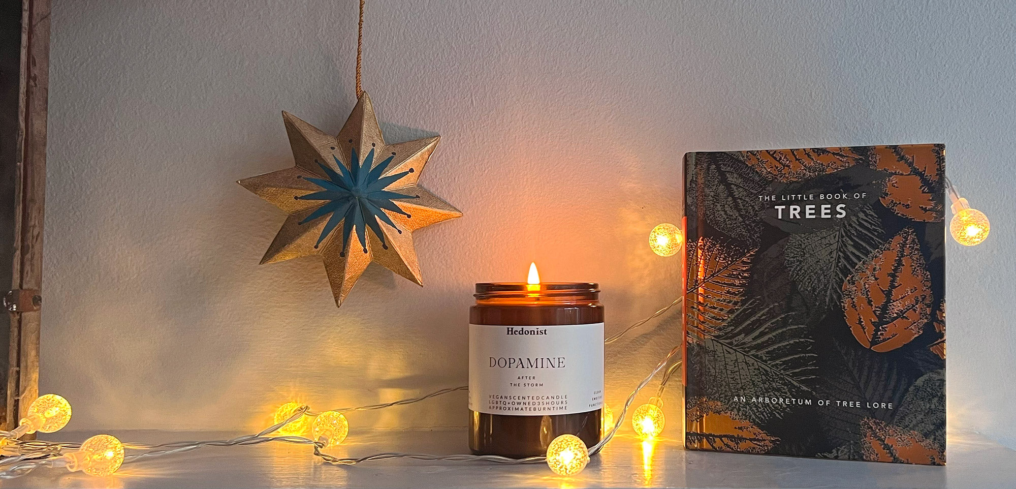 Candle with 'Dopamine' label and decorative book titled 'The Little Book of Trees' on a surface with string lights.