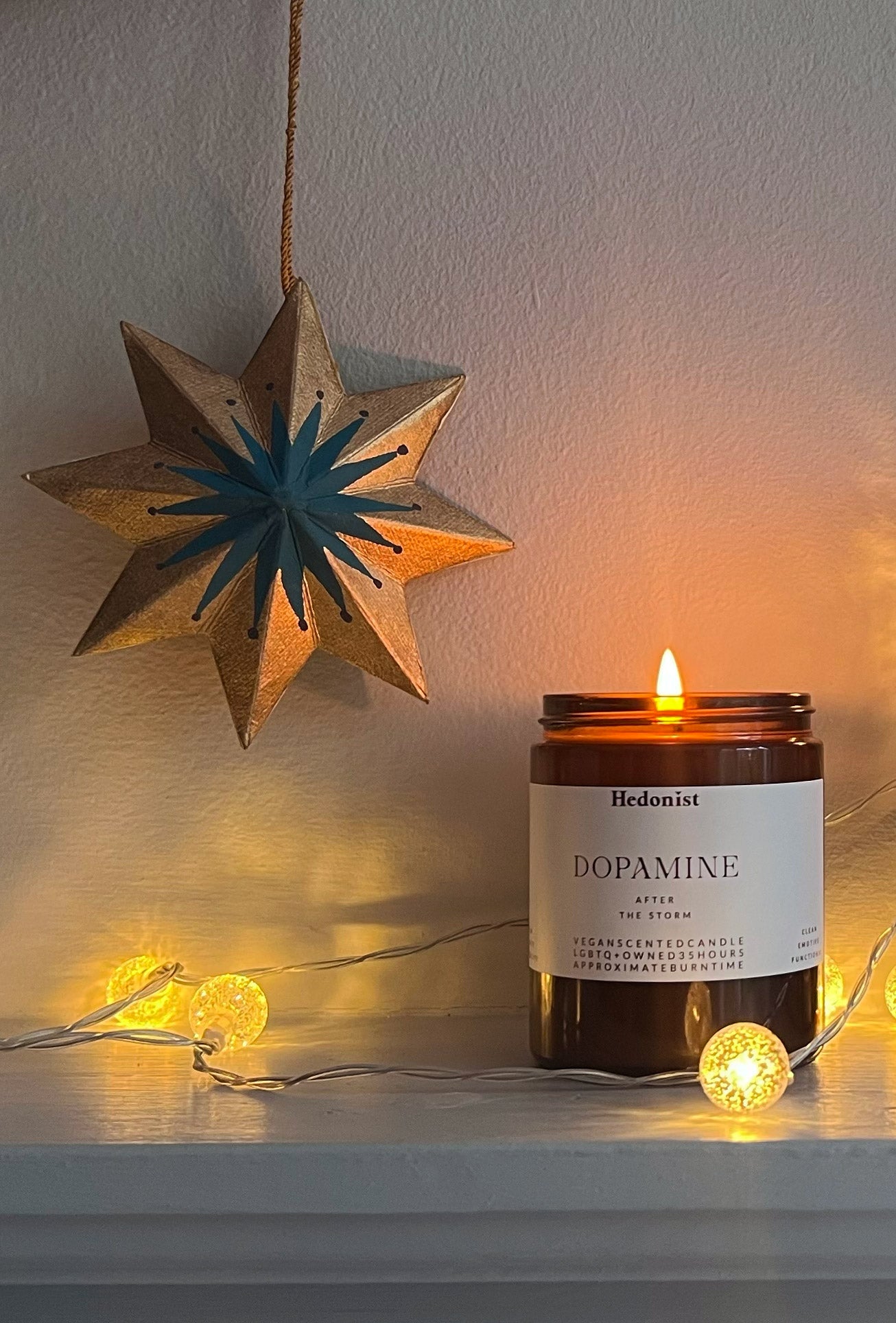 Candle labeled 'Dopamine' by Hedonist with a star-shaped hanging decoration and string lights on a neutral background.